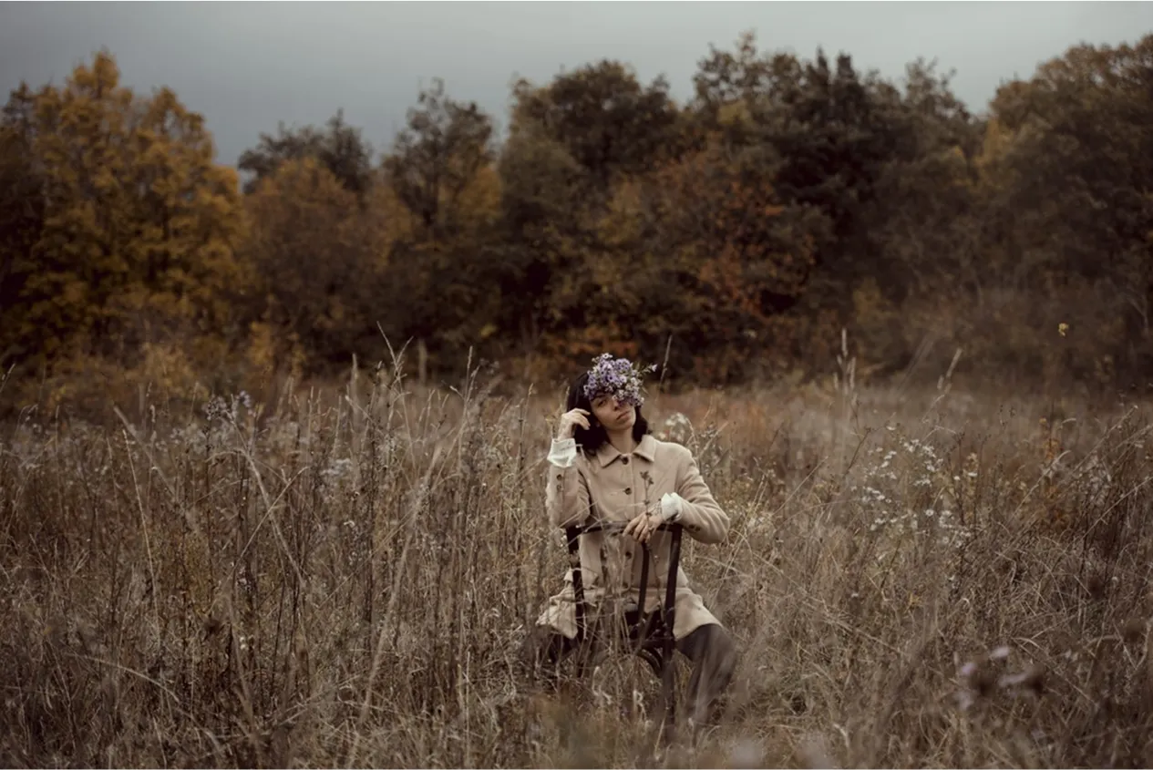 devant une forêt, dans un champ, se trouve une femme assise à l’envers sur une chaise avec un bouquet sur le visage