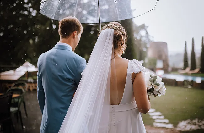 nous voyons de dos un couple de mariés marchant dans un jardin et tenant un parapluie