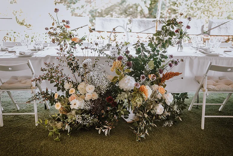 au pied d’une table, lors d’un mariage en extérieur sur une pelouse, se trouve un énorme bouquet de fleurs et de feuilles, composé de blanc, d’orange, de rose et de rose pâle