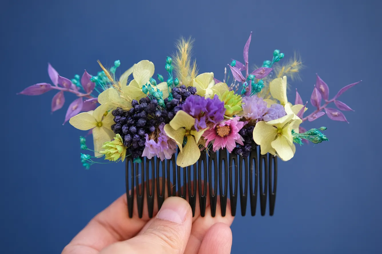 une main tient un peigne avec une composition de fleurs de couleurs vives, comprenant du jaune, du rose, du violet et du bleu