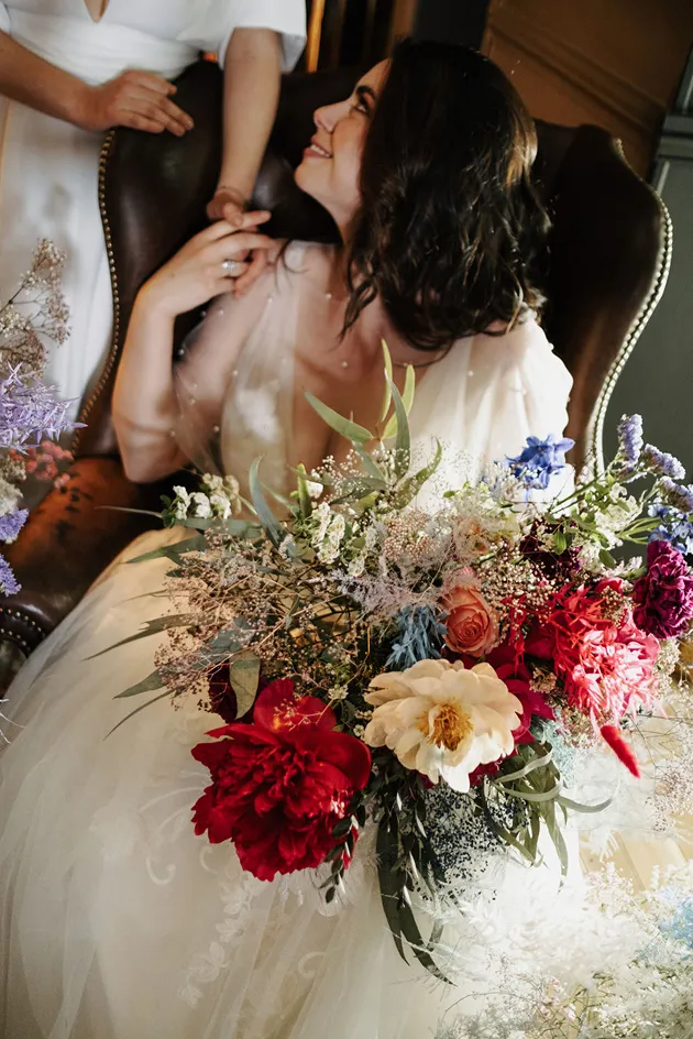 une femme lors d’un mariage, assise sur une chaise et vêtue d’une robe de mariée, tient dans sa main un bouquet de fleurs de couleur rouge, blanc, orange, bleu et rose