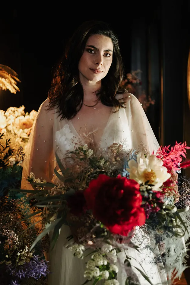 une femme vêtue d’une robe de mariée se tenant debout avec un grand sourire tient dans sa main un énorme bouquet de fleurs composé des couleurs rouge, blanc, rose et violet
