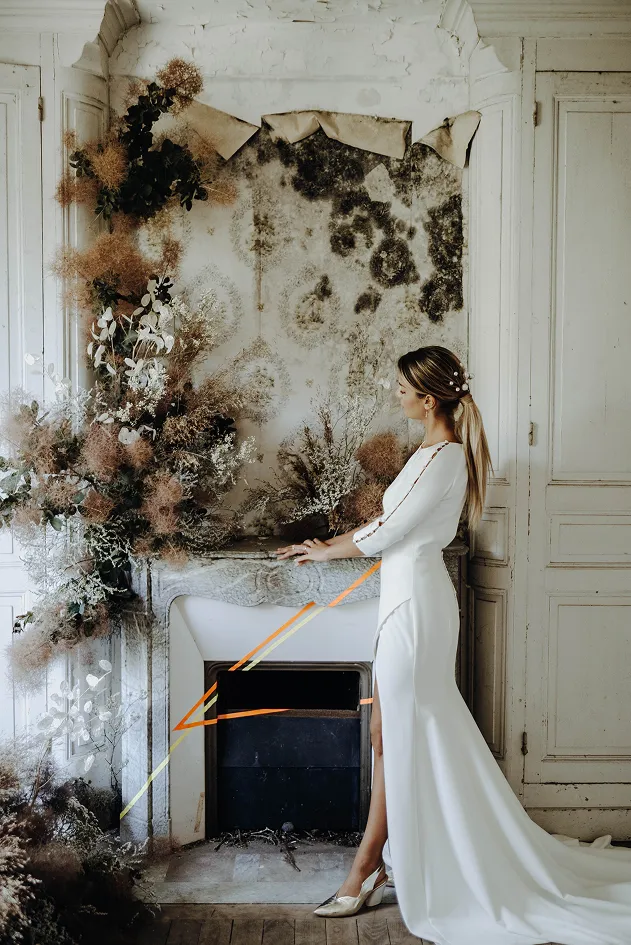 une femme vêtue d’une robe de mariée se tient debout et de profil lors d’un mariage dans une pièce blanche avec des fleurs séchées et un décor vétuste, mise en scène pour le côté artistique