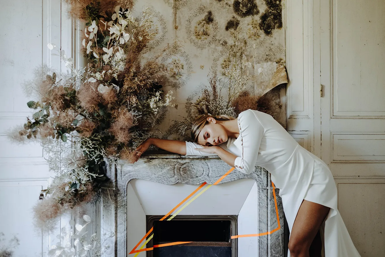 dans un décor vétuste pour un côté artistique se trouve une composition florale de fleurs séchées ainsi qu’une femme vêtue d’une robe de mariée, allongée sur une cheminée