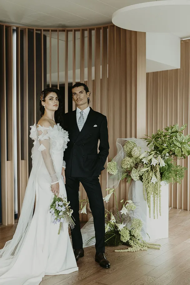 dans un décor contemporain se trouve un couple de mariés avec la mariée, vêtue d’une robe, tenant un bouquet de fleurs dans sa main droite, et un homme en costume