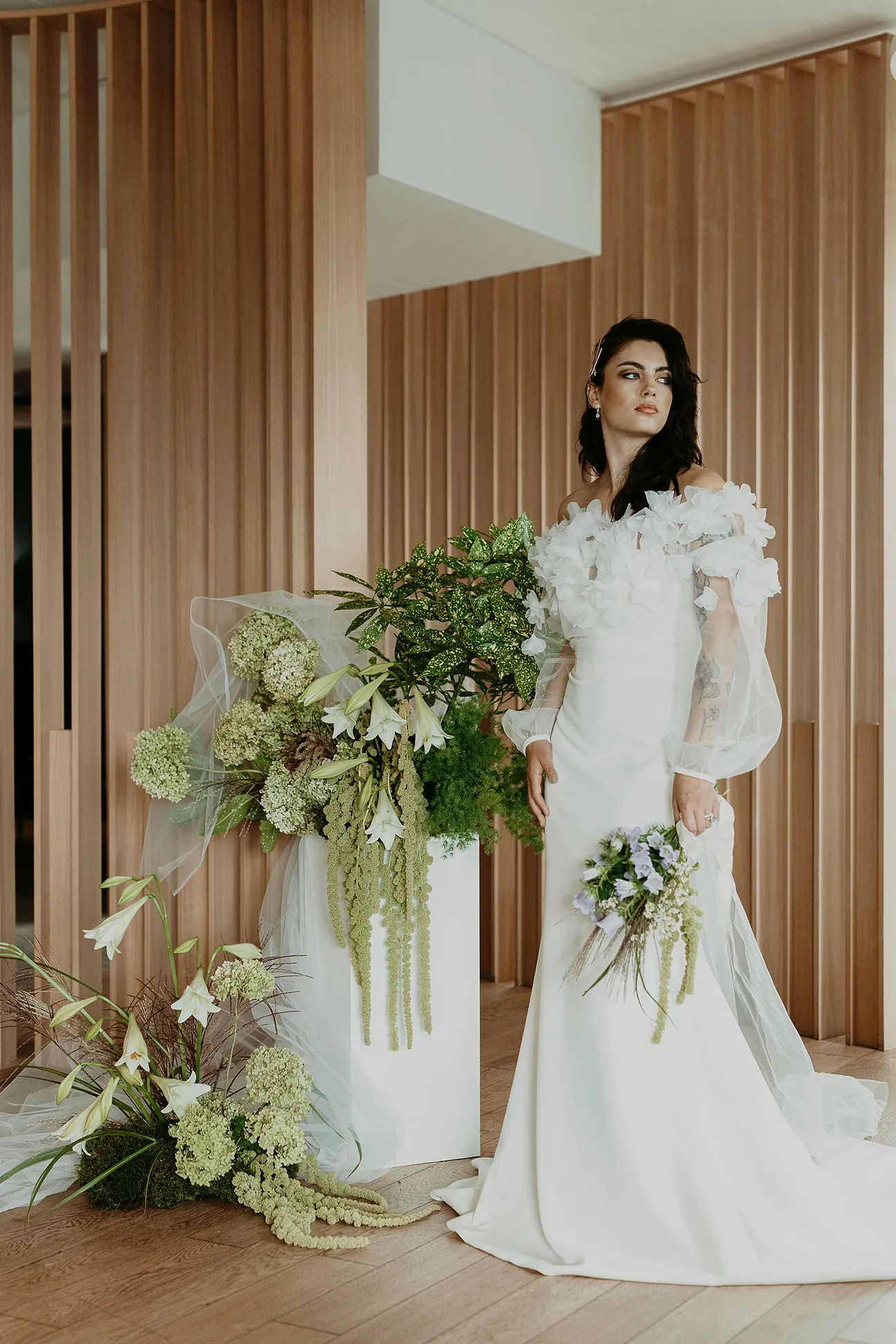 un décor contemporain avec des couleurs blanches et bois se trouve une femme vêtue d’une robe de mariée, se tenant de face et tenant dans sa main gauche un bouquet de fleurs ainsi que beaucoup de fleurs autour d’elle, principalement vertes et blanches