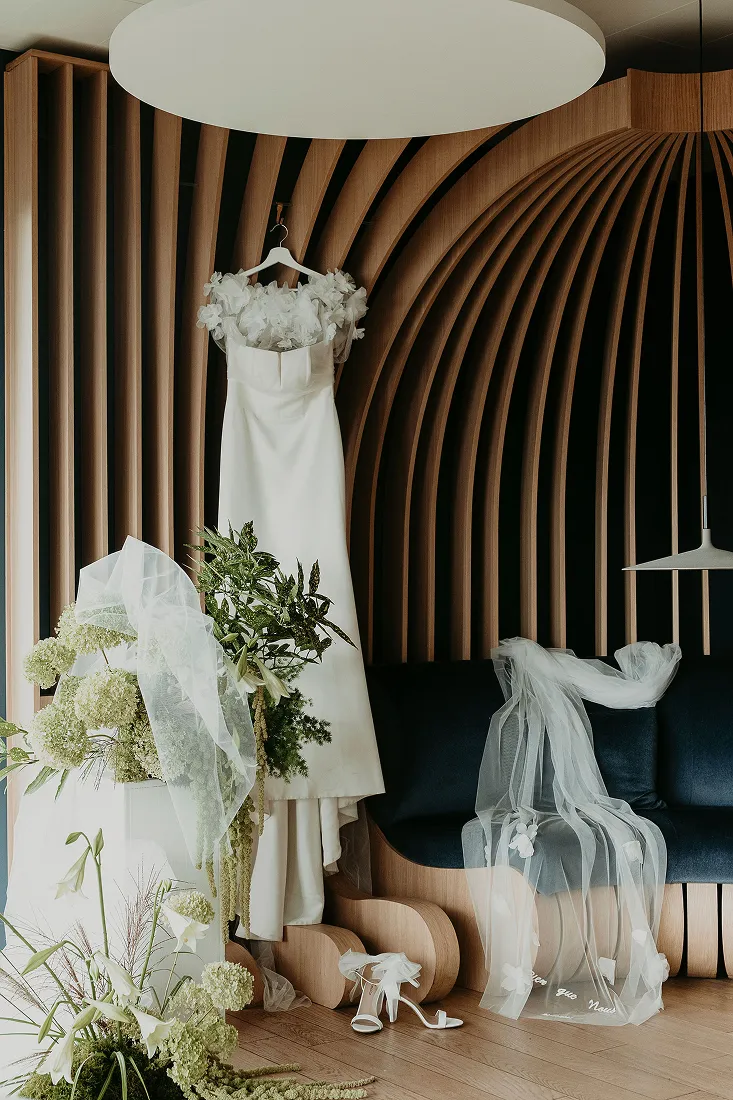 dans un décor contemporain avec les couleurs bois clair se trouve une robe de mariée suspendue à un cintre ainsi que des fleurs de couleur blanche et verte