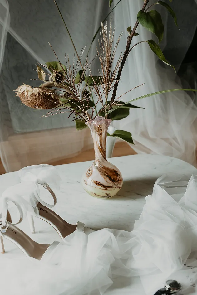 sur une table blanche se trouve un vase avec des fleurs marron clair et vertes, ainsi que des chaussures à talons et un voile blanc