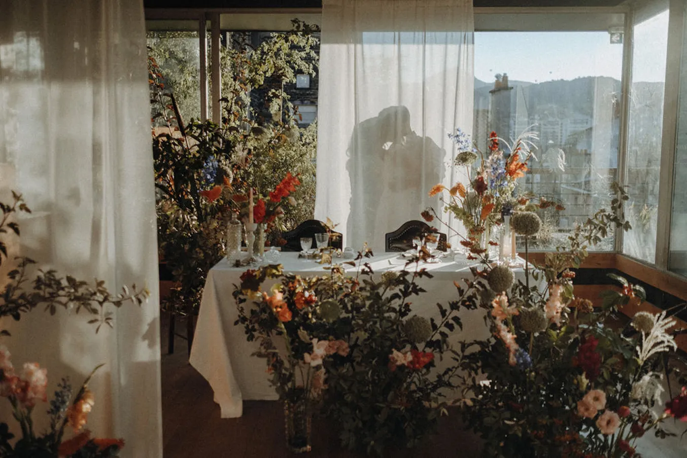 dans un décor rempli de fleurs derrière un voile blanc se trouve un couple de mariés s’embrassant