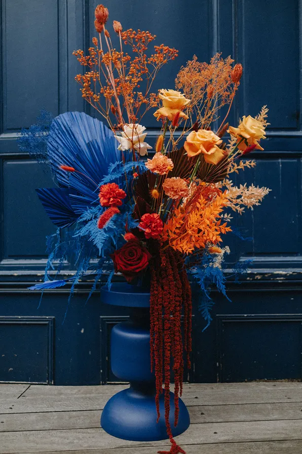 sur fond bleu, dans un vase bleu se trouvent des fleurs composées des couleurs bleu, rouge et orange
