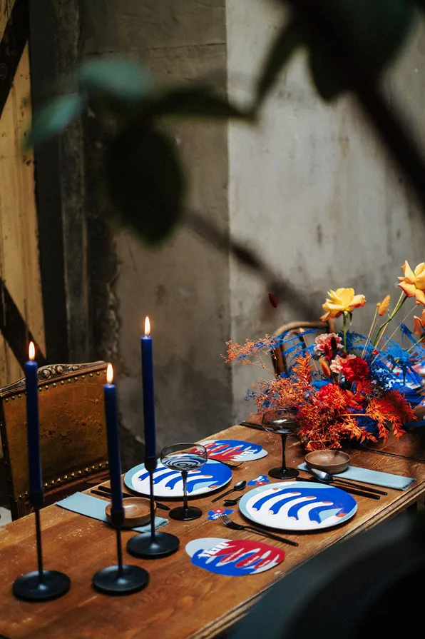 sur une table en bois, trois bougies bleues et grandes allumées ainsi qu’une table dressée avec des fleurs de couleur rouge, orange, bleu et jaune