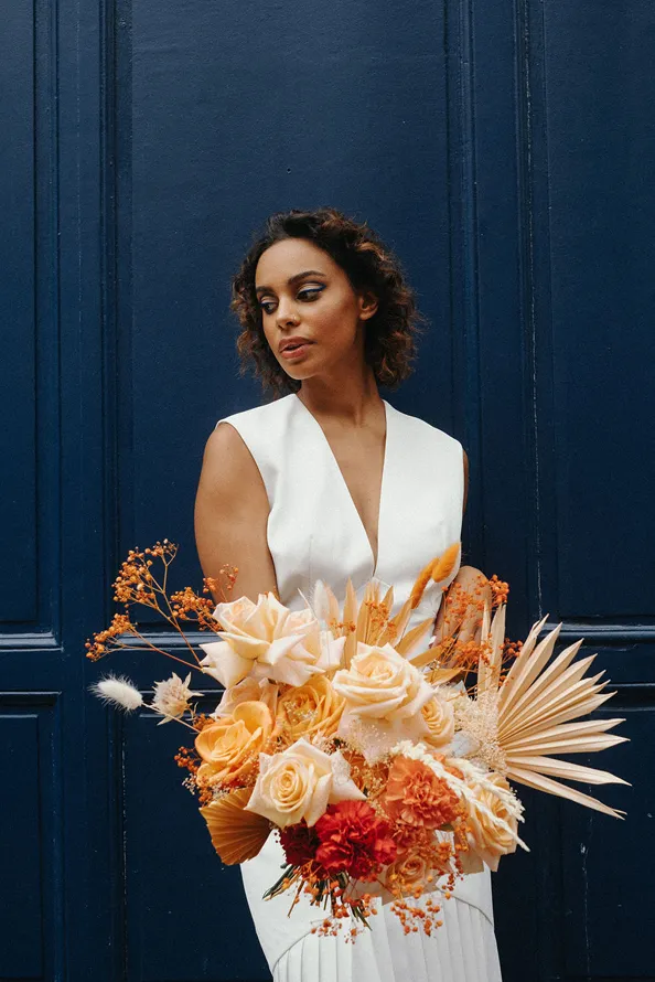 une femme vêtue d’une robe de mariée tient dans ses mains un bouquet de fleurs de couleur pâle, avec un fond bleu derrière elle