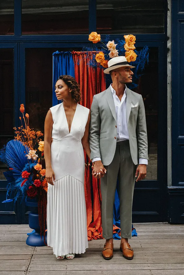 en premier plan se trouve un couple de mariés se tenant la main, et derrière se dressent des fleurs de couleur jaune, bleu et rouge