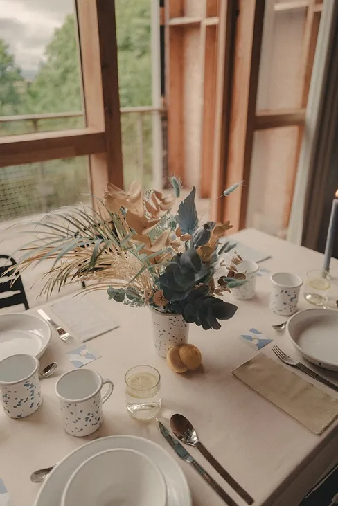 sur une table blanche dressée se trouve un petit vase blanc avec des fleurs de couleur bleue, verte et dorée