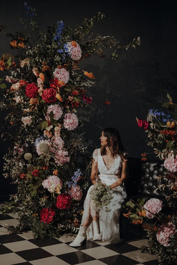 entourée de diverses fleurs de diverses couleurs se trouve une femme vêtue d’une robe de mariée, assise et tenant un bouquet de fleurs blanches dans ses mains