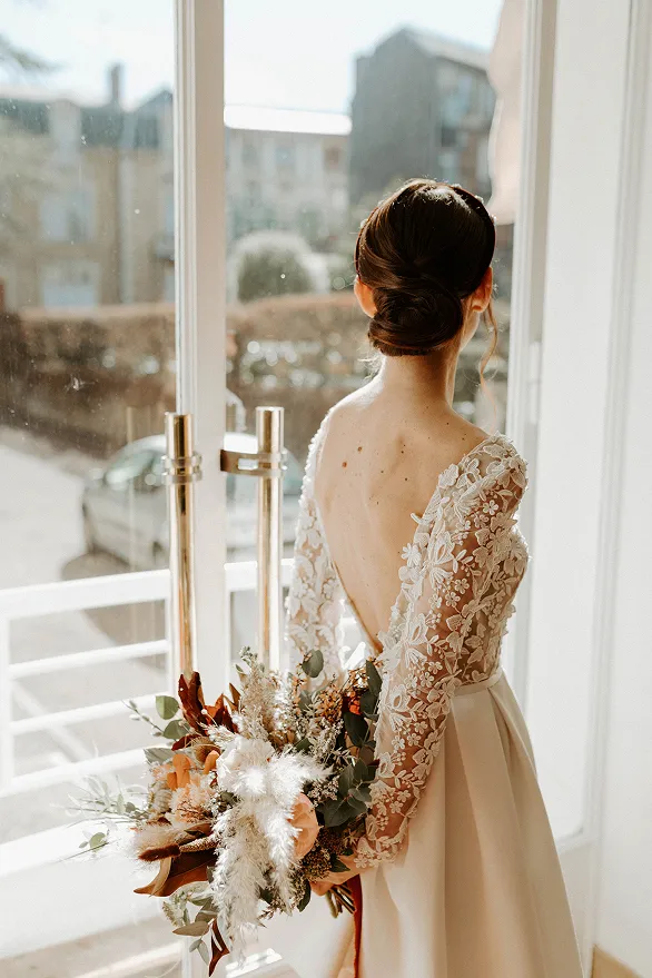 une femme de dos, vêtue d’une robe de mariée, tient dans ses mains un bouquet de fleurs et regarde par la vitre